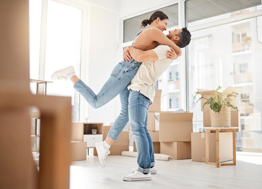 Casal feliz comemorando mudança em apartamento novo com caixas de mudança no fundo.