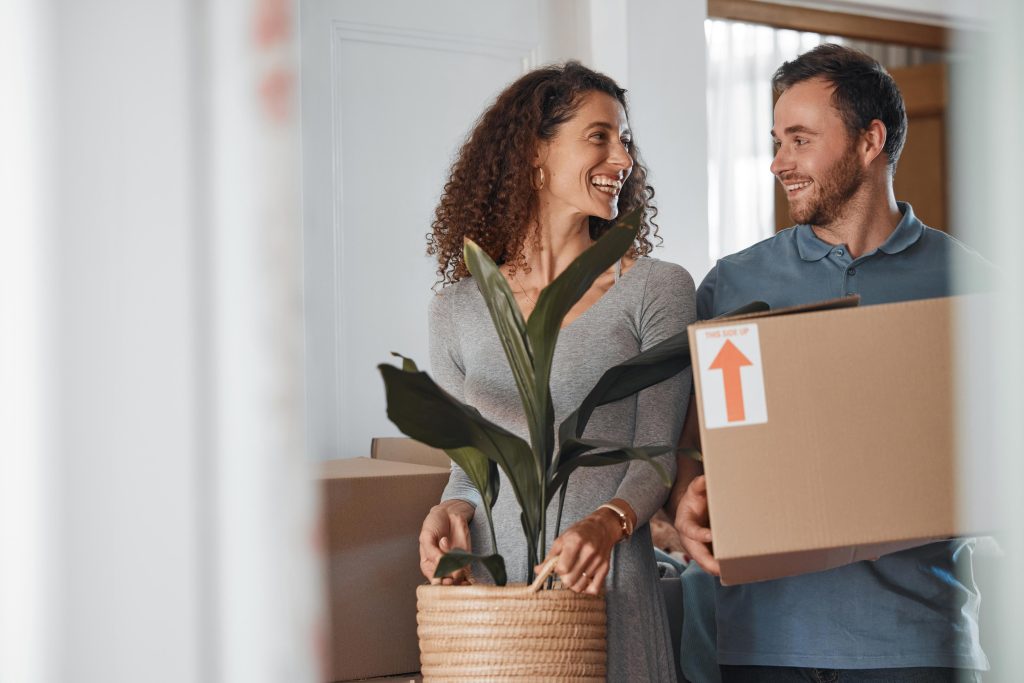 Casal sorridente carregando planta e caixa durante mudança para novo apartamento